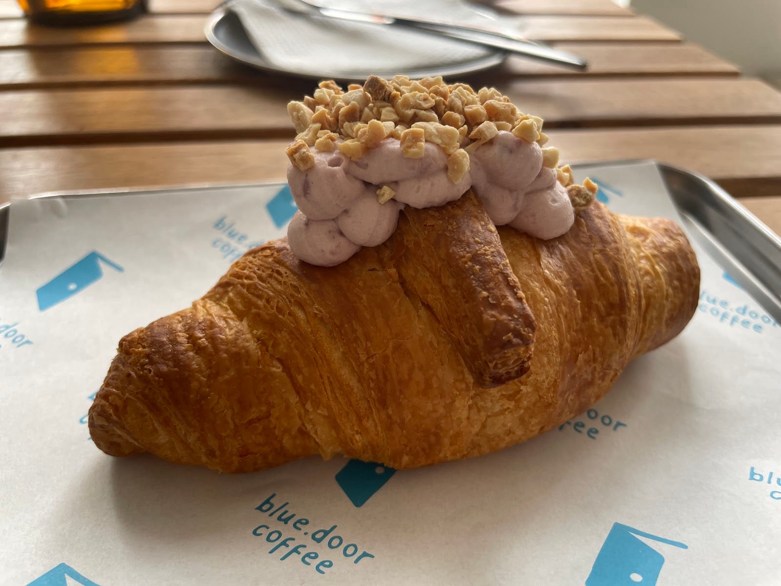 A dessert croissant topped with cream and crushed nuts, served on a Blue Door Coffee branded paper tray